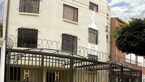 A building with a white facade, barred windows, and a green gate. - Apartamentos Los Alcázares (Bogotá)