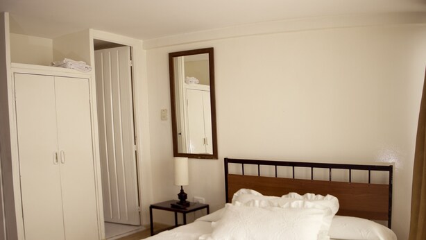 A neatly made bed with white linens in a room with wooden flooring, a white wardrobe, and a small bedside table with a lamp. - Apartamentos Los Alcázares (Bogotá)