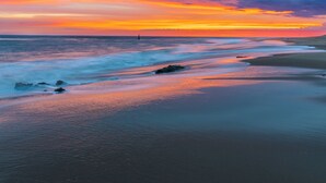 A beach with a sunset, waves, and a distant lighthouse.