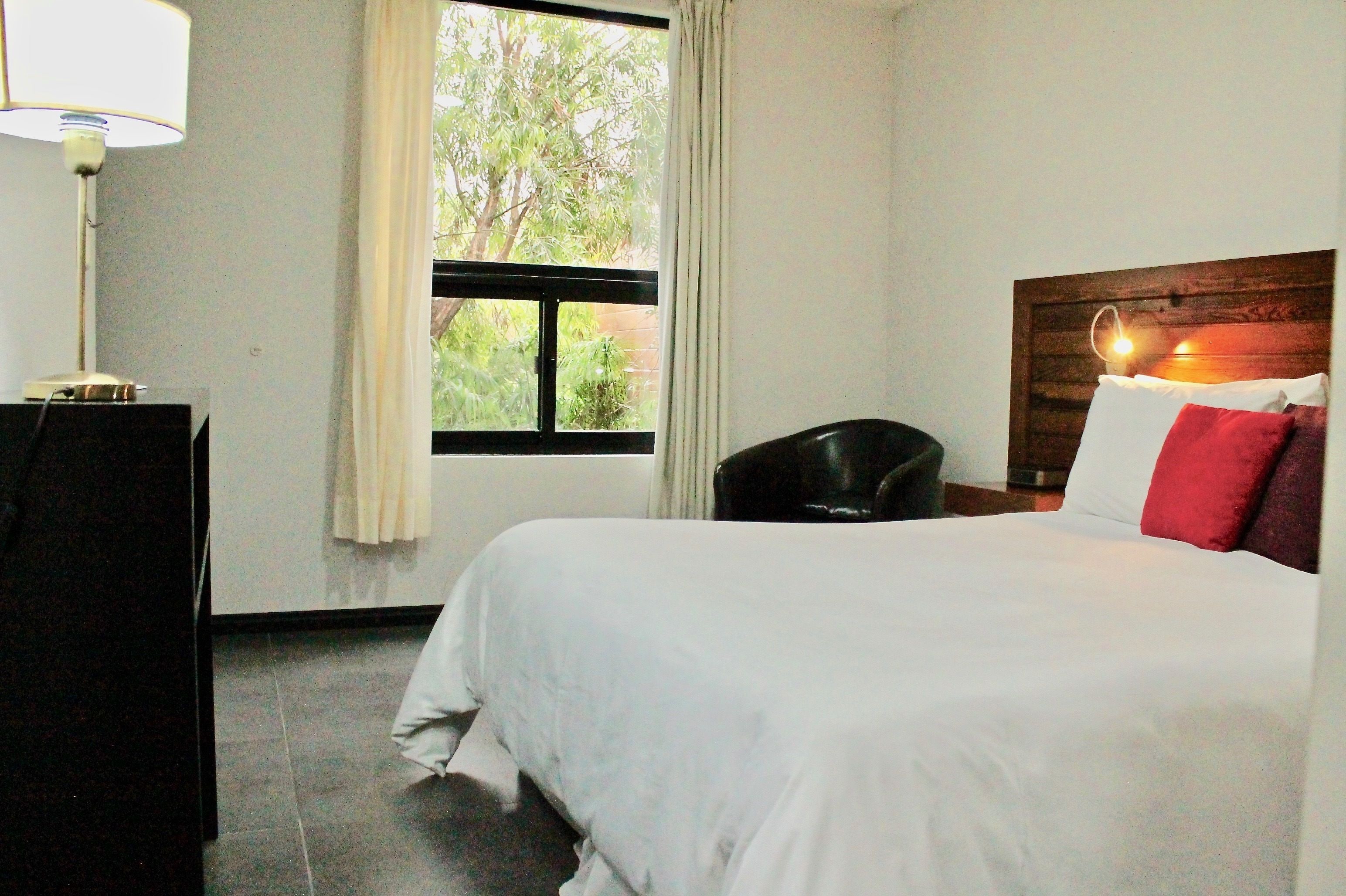 A hotel room with a bed, a desk, a chair, a window with curtains, and a lamp.