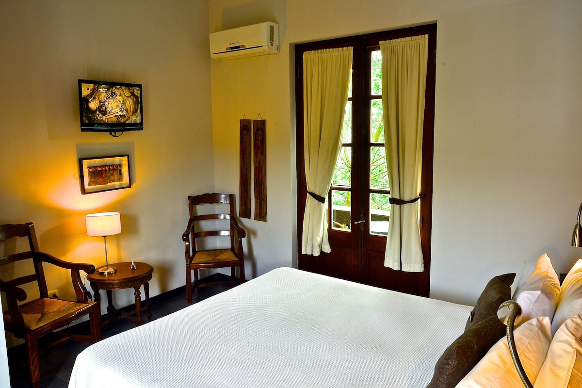 Habitación de hotel con cama, dos sillas, una mesa con lámpara, un cuadro y una ventana con cortinas.