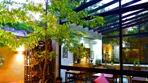 An outdoor dining area with wooden tables and chairs, surrounded by greenery and a glass wall.