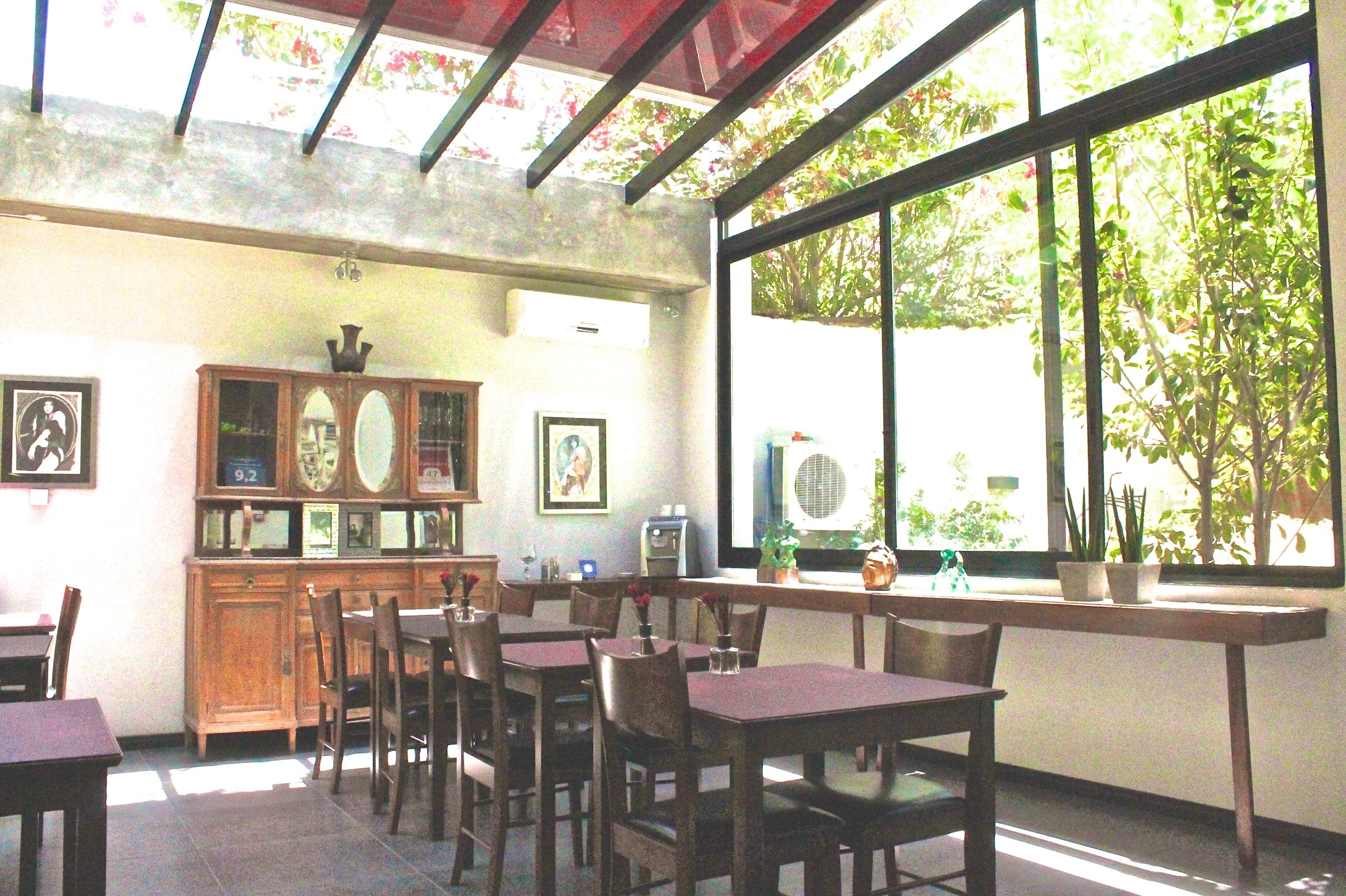 A restaurant with wooden tables and chairs, a wooden cabinet with framed pictures, and large windows with a view of greenery outside.