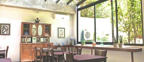 A restaurant with wooden tables and chairs, a wooden cabinet with framed pictures, and large windows with a view of greenery outside.