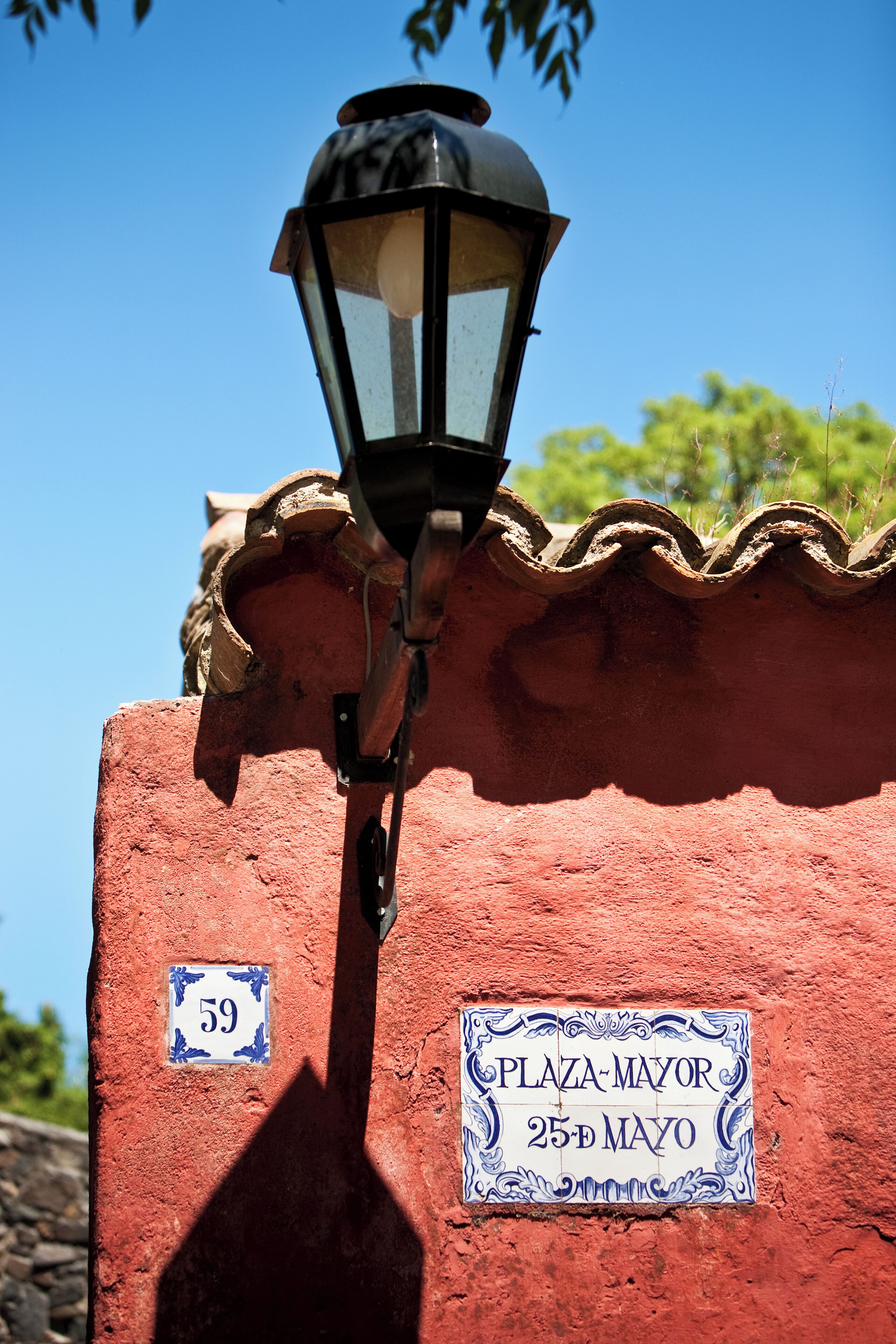 A street lamp attached to a building with a red facade and a sign reading "Plaza-Major 25th Mayo".
