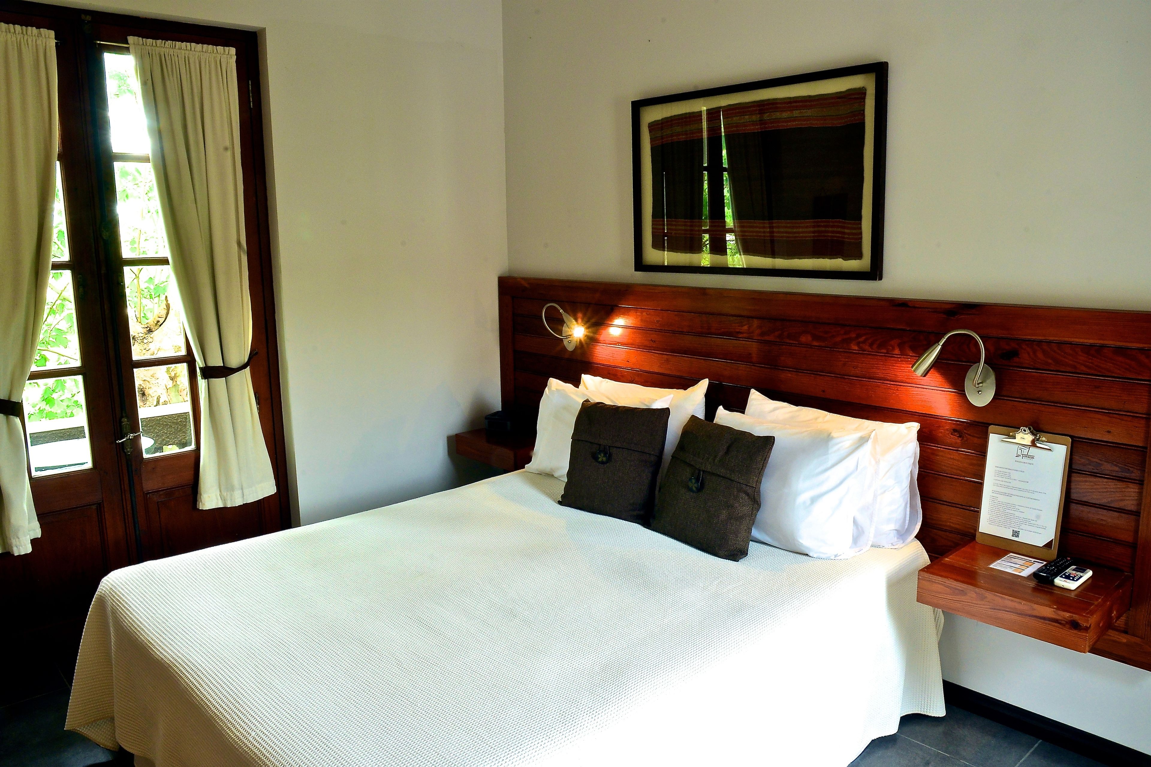 A neatly made bed with white and brown bedding, a wooden headboard, a bedside table with a lamp, and a framed picture on the wall.