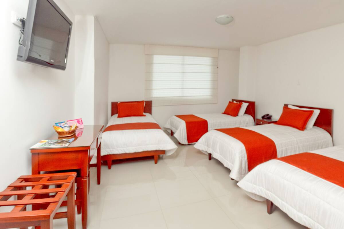 A hotel room with four beds, a TV, a desk, and a window with blinds.