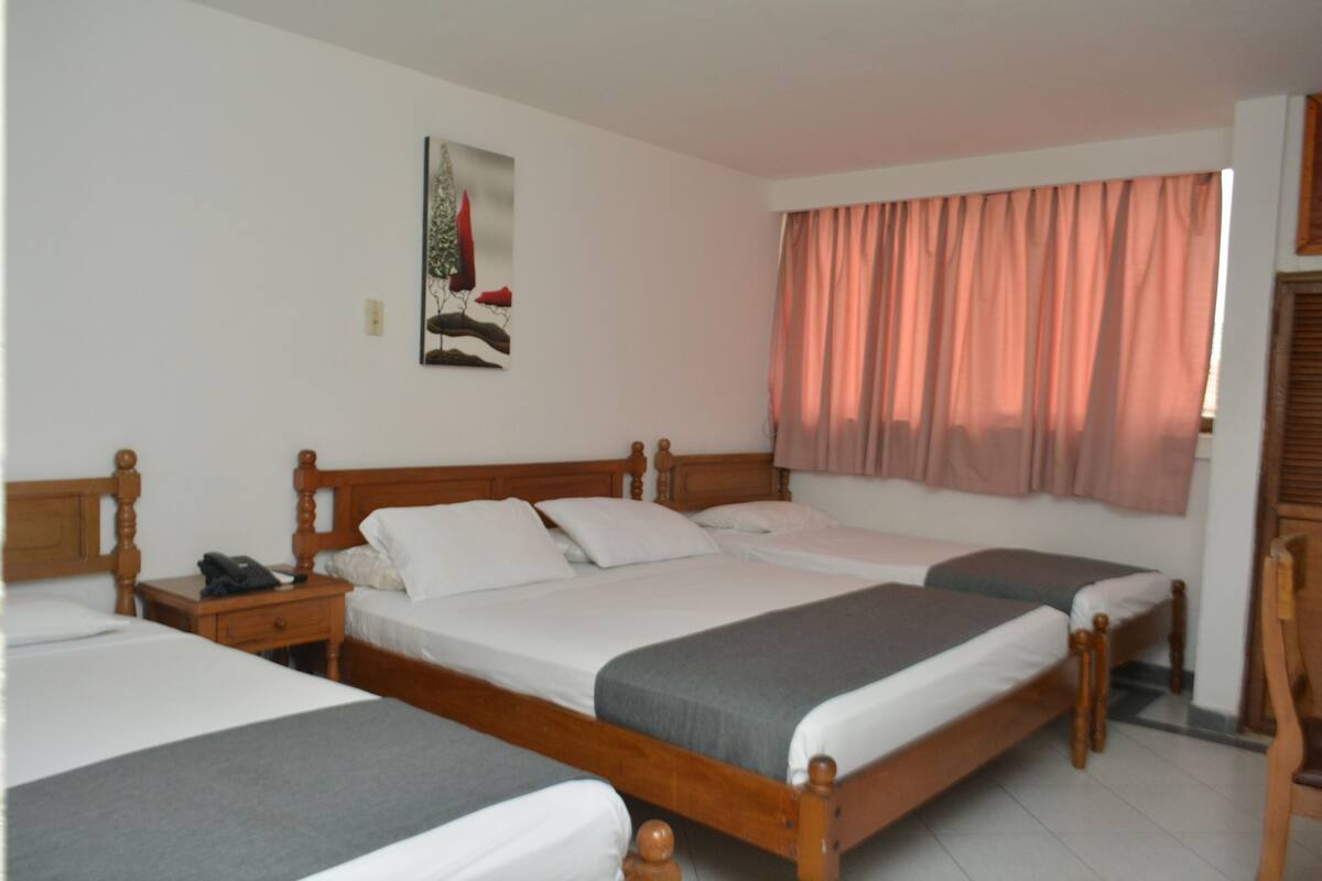 Una habitación de hotel con tres camas de madera, una mesita de noche con teléfono, un cuadro en la pared y una ventana con cortinas rosadas.