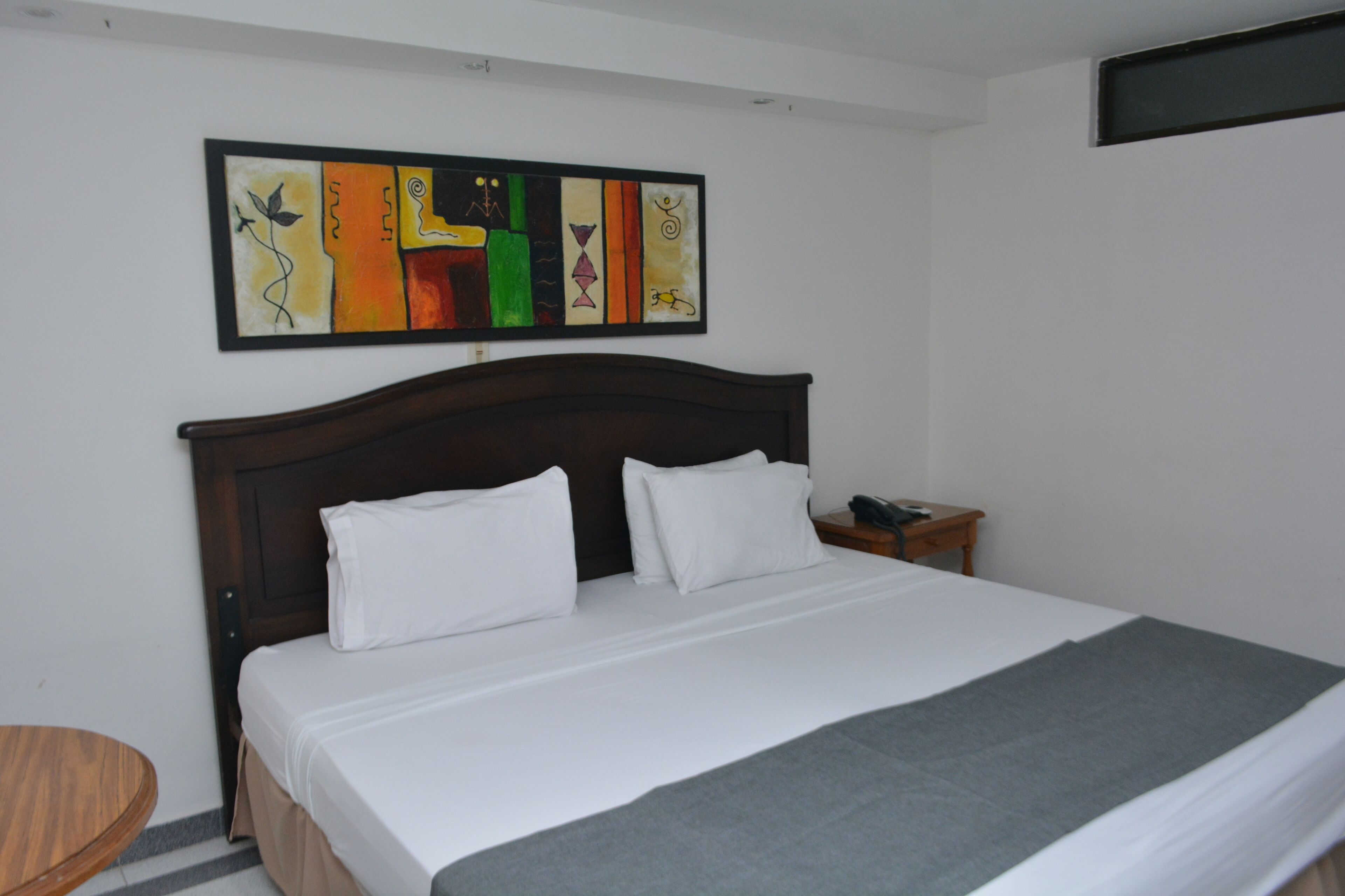 Een net opgemaakt bed met witte lakens en een grijze plaid, een houten nachtkastje met een telefoon, en een kleurrijk abstract schilderij aan de muur.