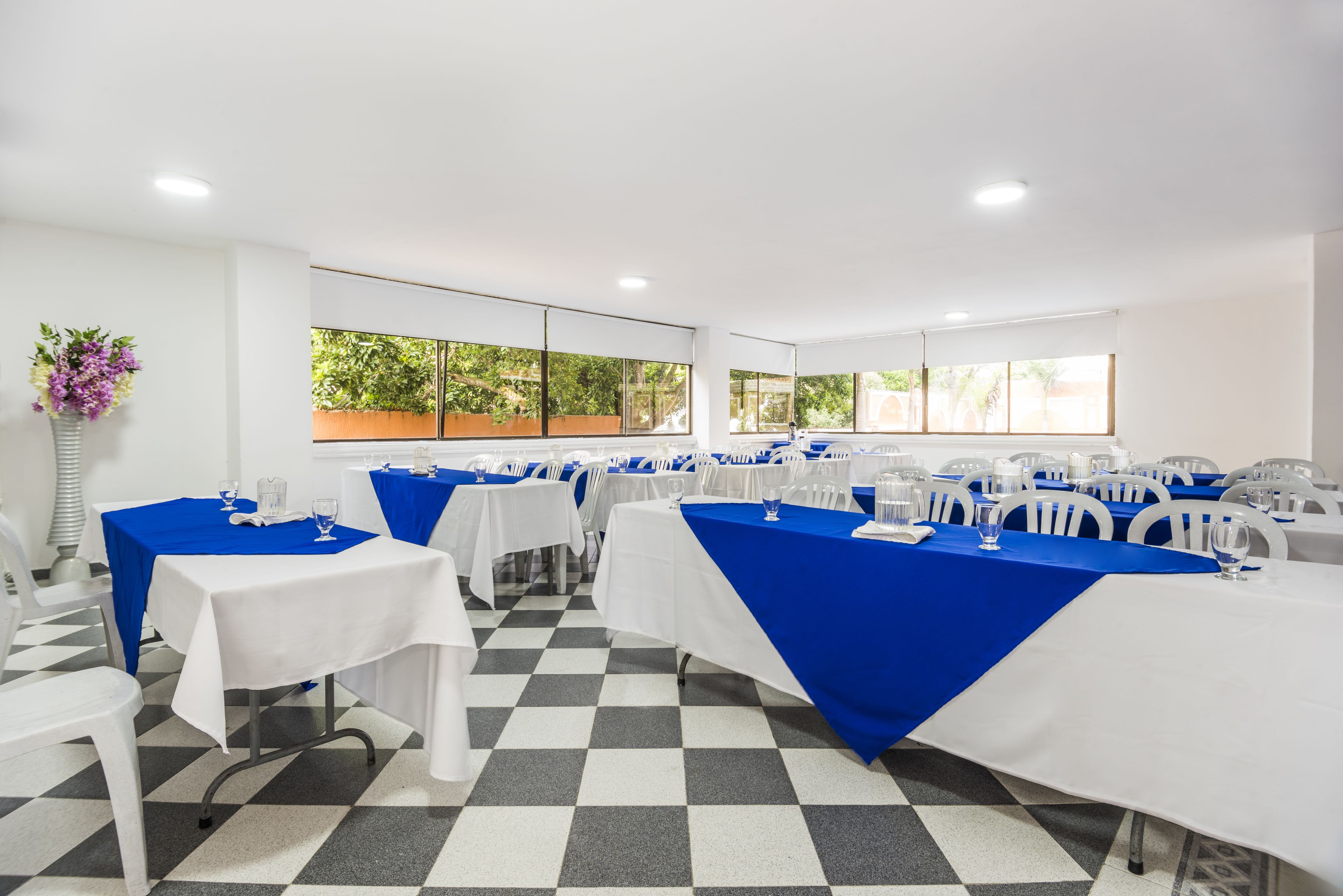 Un salón de conferencias con mesas dispuestas para una reunión, ventanas amplias con follaje exterior y un suelo a cuadros.