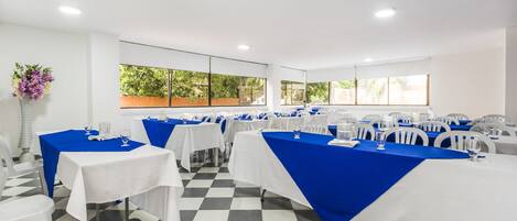 Un salón de conferencias con mesas dispuestas para una reunión, ventanas amplias con follaje exterior y un suelo a cuadros.