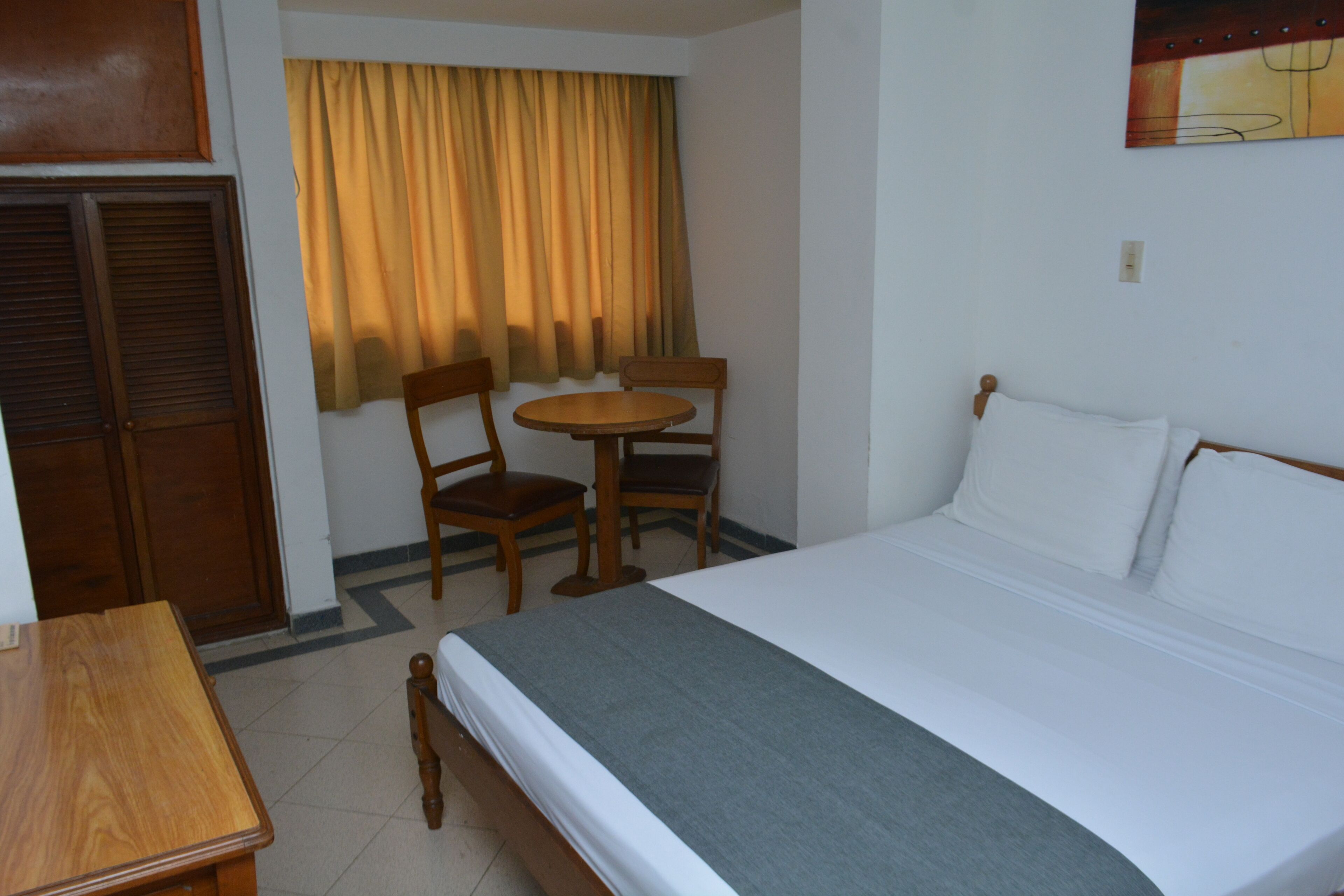 A hotel room with a bed, a wooden table, two chairs, and a window with curtains.