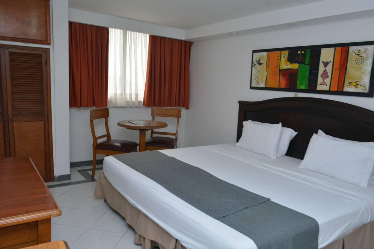 Una habitación de hotel con cama, una mesa de madera, una silla y una ventana con cortinas.