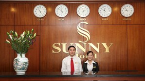 A reception desk with clocks displaying times for Tokyo, Seoul, Hanoi, Paris, and London. There is a vase with flowers and the word "SUNNY" on the wall. - Sunny 3 Hotel Hanoi (Hanoi)