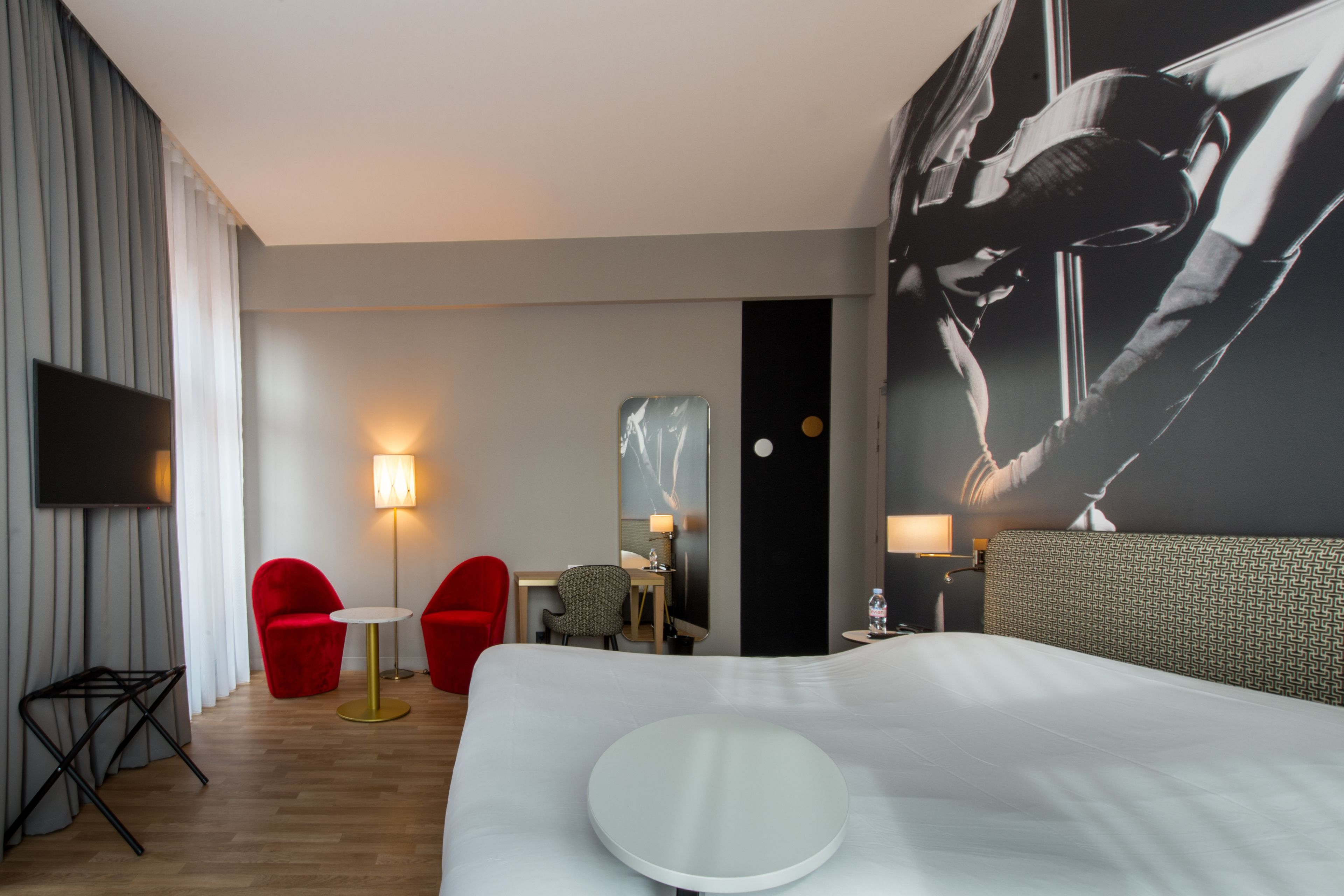 A hotel room with a large bed, a flat-screen TV, two red chairs, a small round table, and a wall-mounted mirror.