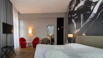 A hotel room with a large bed, a flat-screen TV, two red chairs, a small round table, and a wall-mounted mirror.