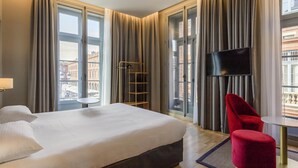 A hotel room with a bed, a TV, a red chair, and a view of buildings through large windows. - Ibis Styles Toulouse Capitole (Toulouse)