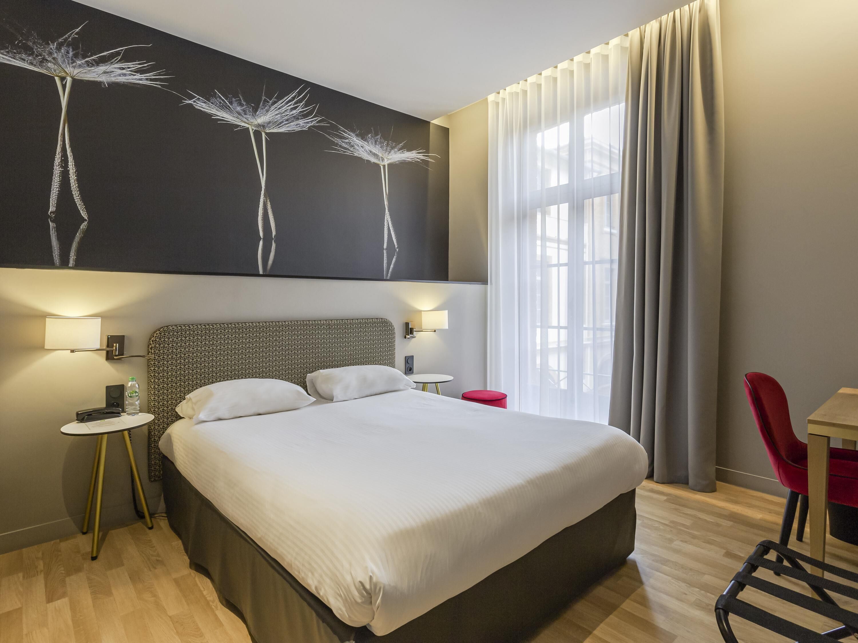 Una habitación de hotel moderna con una cama grande, dos mesitas de noche, una silla roja, un escritorio y un ventanal con cortinas.