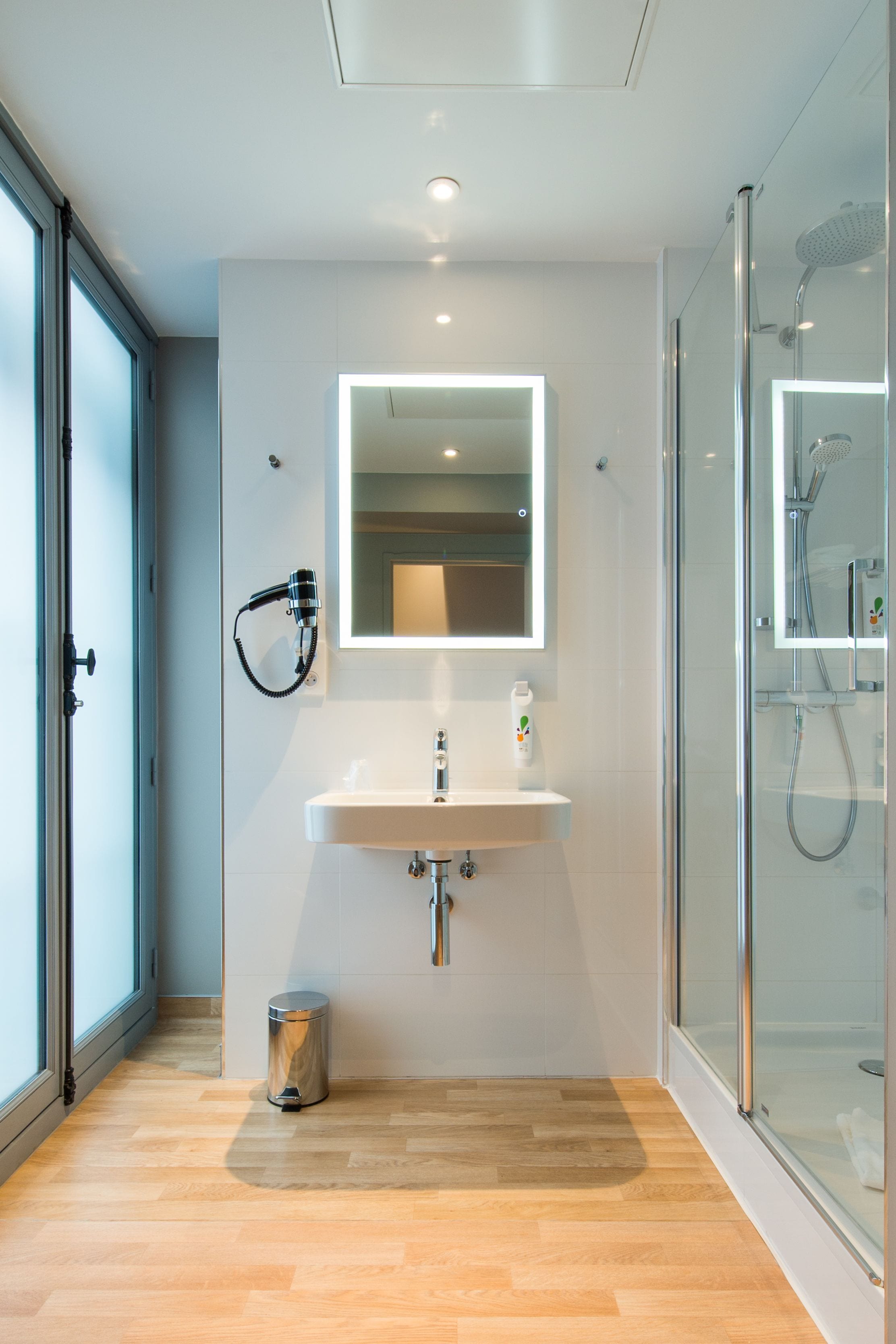 Een moderne badkamer met een douchecabine van glas, een wit wastafelmeubel met spiegel en een kleine prullenbak.