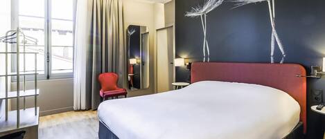 A hotel room with a large bed, a red chair, a mirror, and a wall art piece.