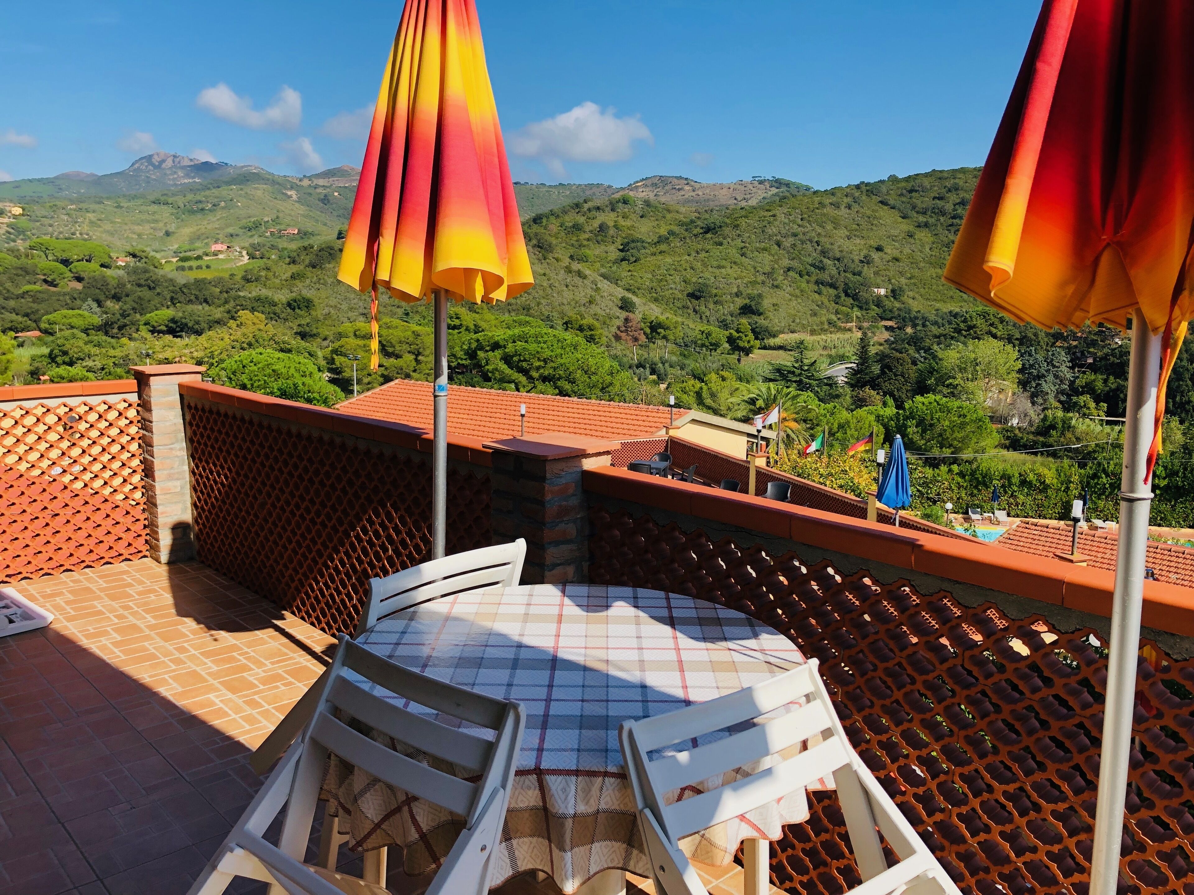 A terrace with a table and chairs, a view of green hills, and a red-tiled roof.