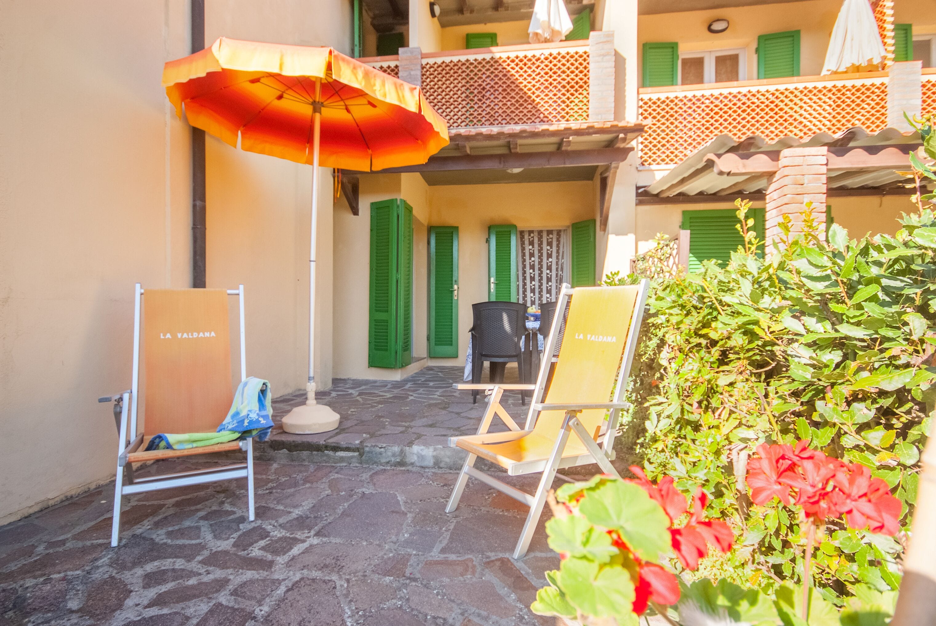 An outdoor seating area with two lounge chairs and an umbrella, a stone-paved patio, and a balcony with green shutters.