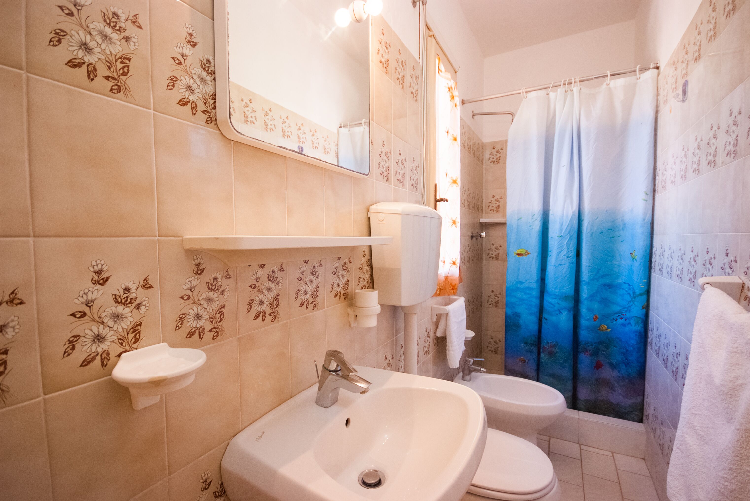 Une salle de bain avec un lavabo, deux toilettes, un miroir et un rideau de douche orné de motifs de poissons.