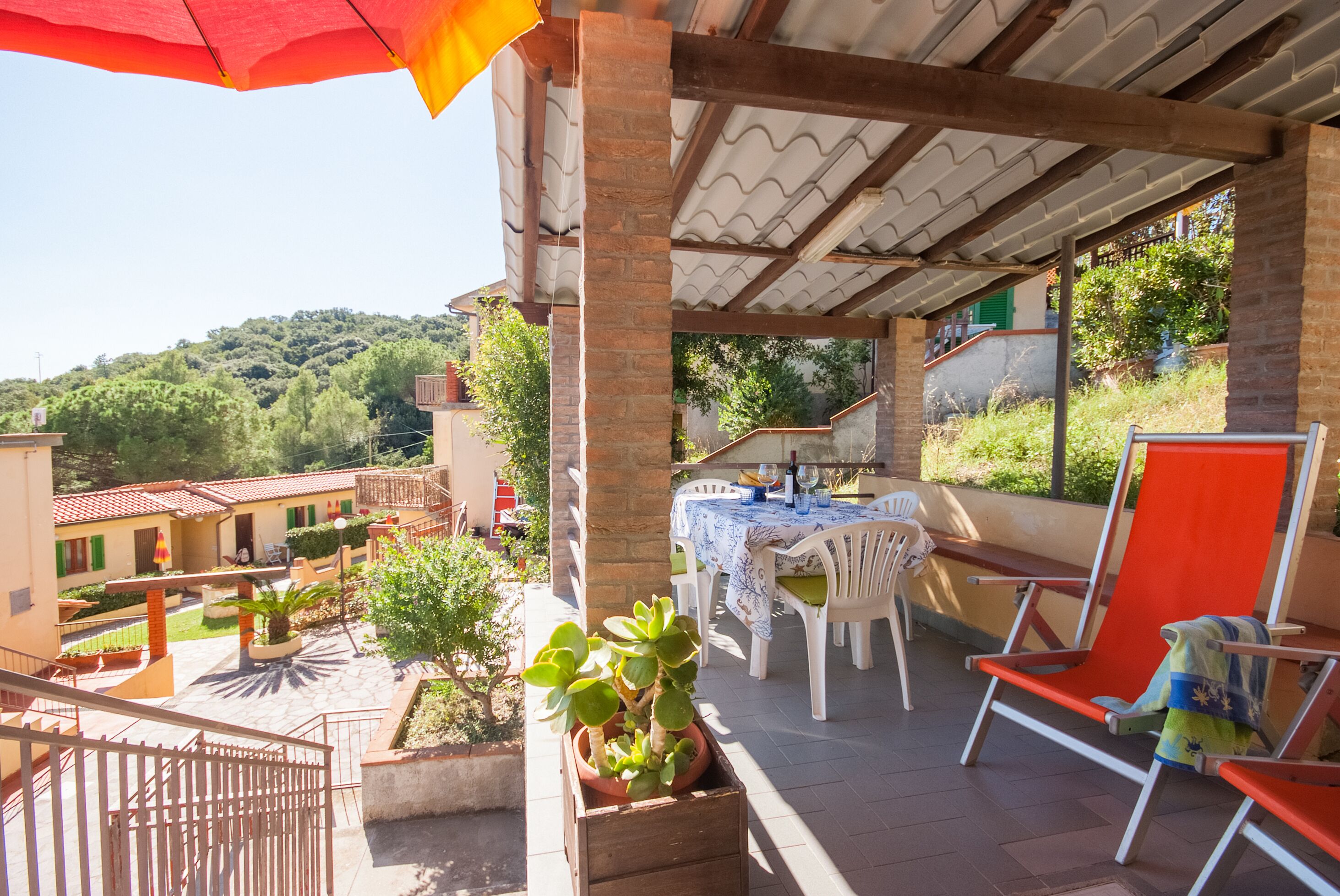 Eine überdachte Terrasse im Freien mit Tisch und Stühlen, Blumentöpfen und Blick auf die Umgebung.
