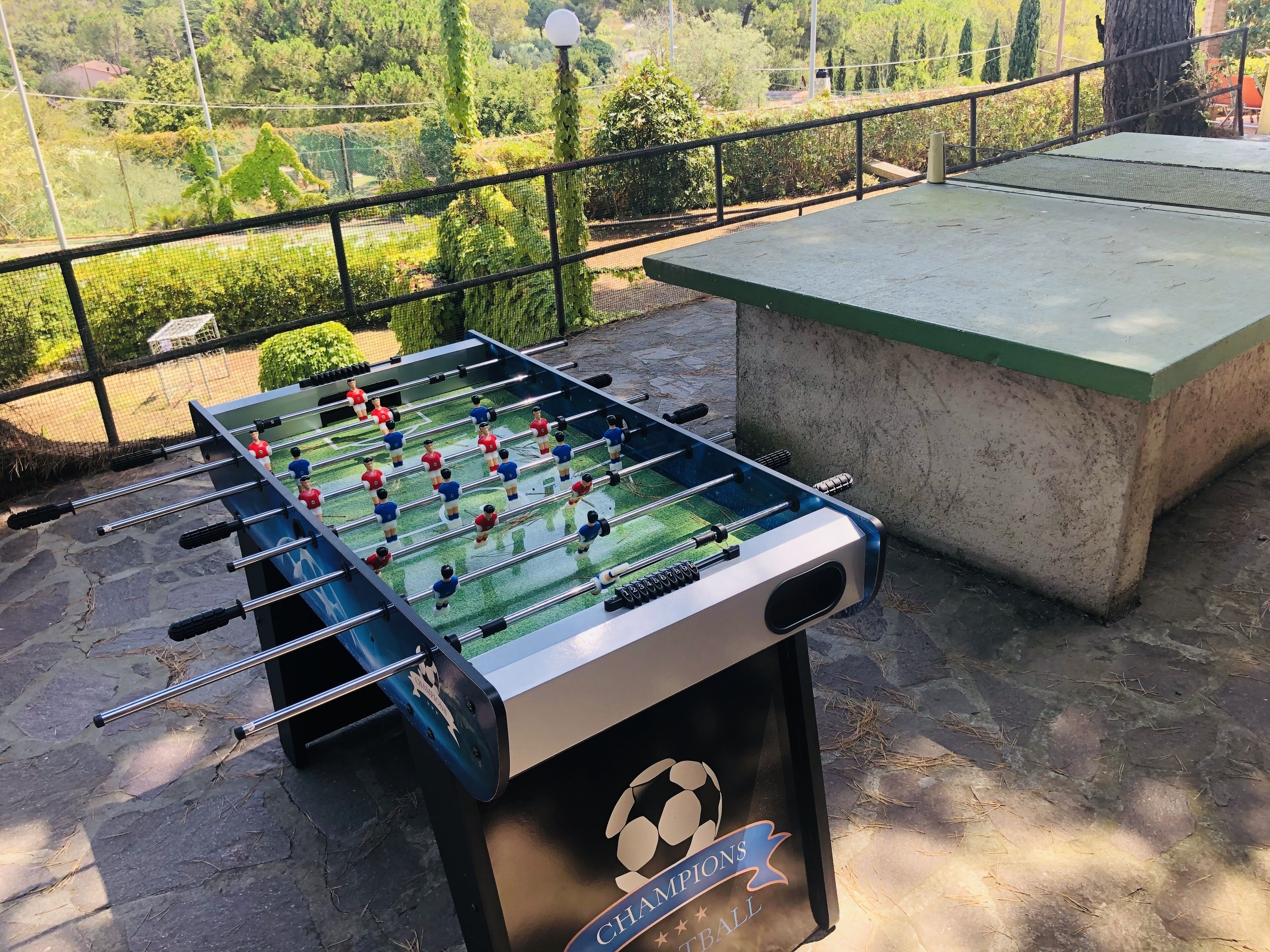 A foosball table with a soccer ball logo on the side, placed on a stone surface with a green table nearby.