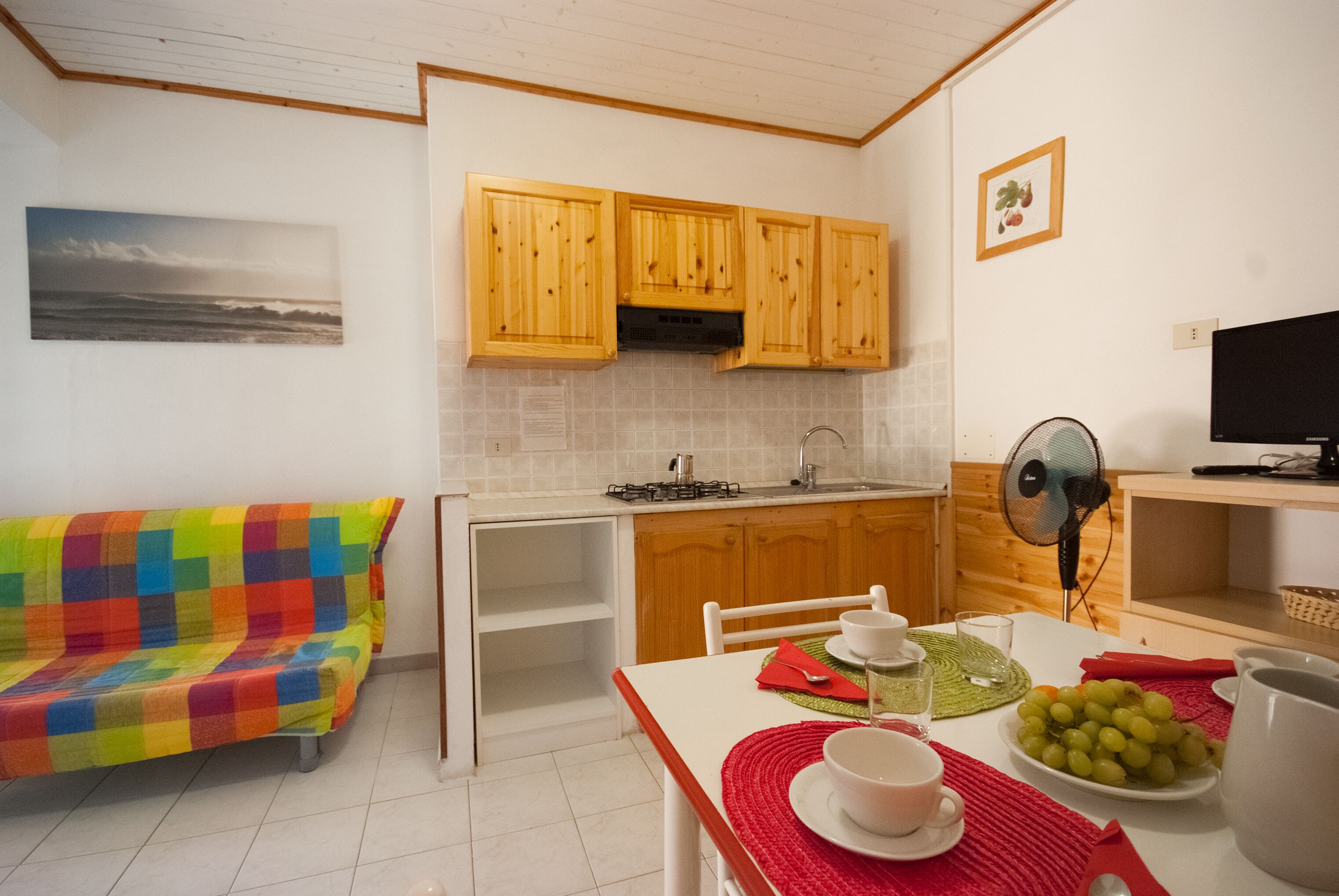 A compact kitchen with wooden cabinets, a dining table with a red placemat, and a colorful sofa.