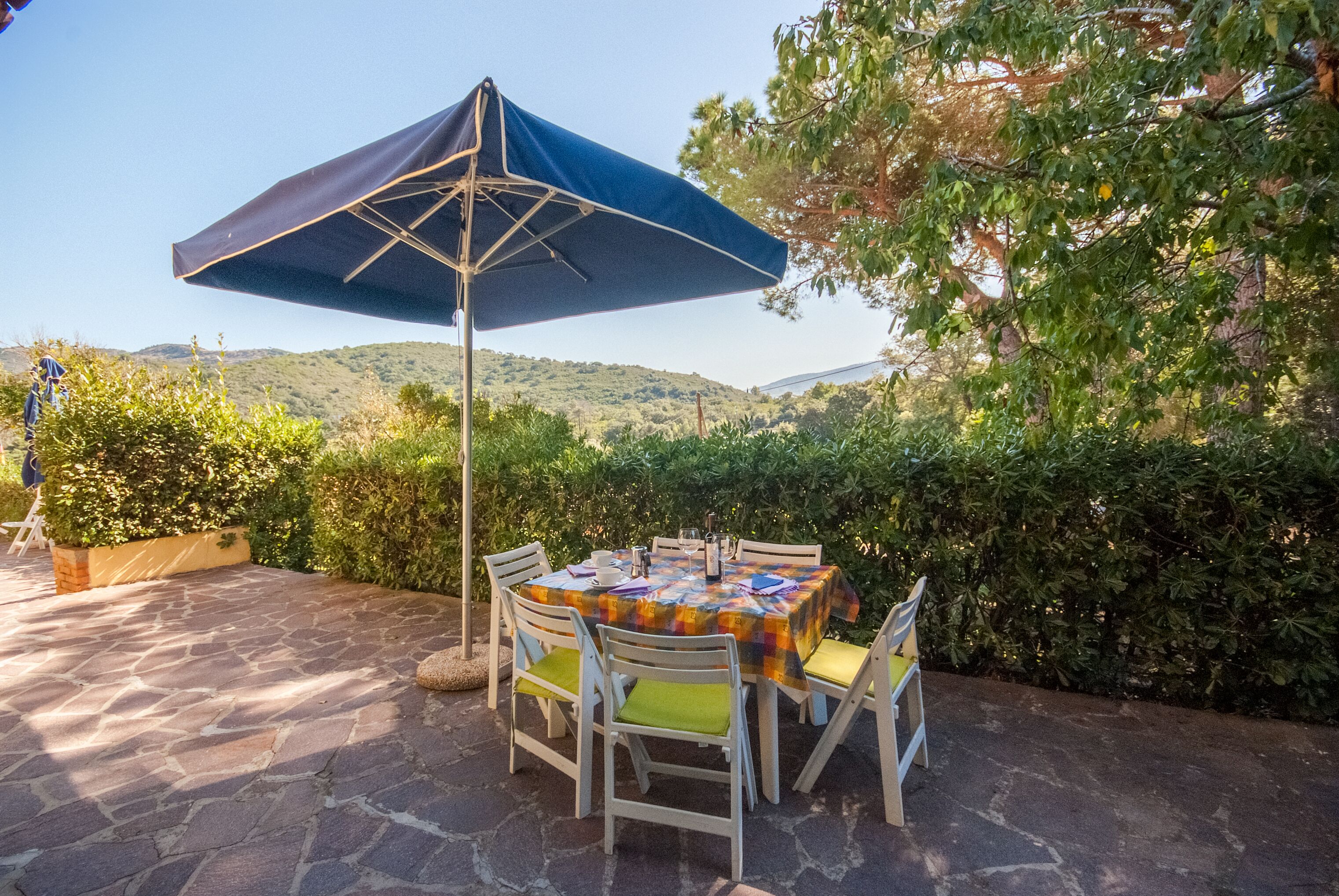 Eine Terrasse im Freien mit Tisch und Stühlen, einem blauen Sonnenschirm und Blick auf Hügel im Hintergrund.