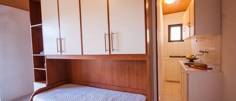Ein Schlafzimmer mit einem Holzbett, weißen Schränken und einer kleinen Küchenzeile mit Fenster.