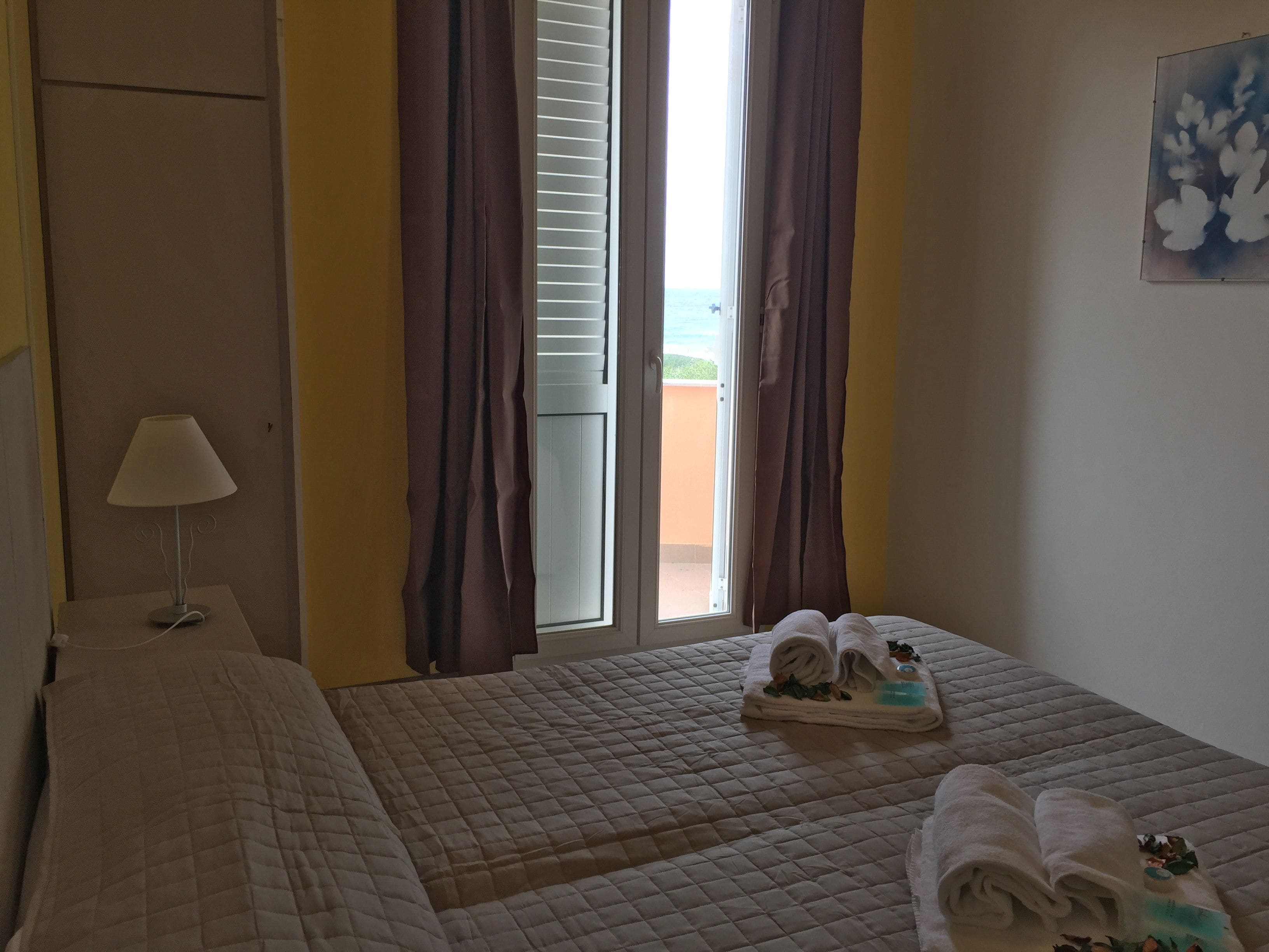 A hotel room with a bed, a nightstand with a lamp, a window with curtains, and a view of the outside.