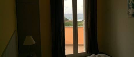 A hotel room with a bed, a nightstand, a lamp, and a view of the sea through a sliding door.
