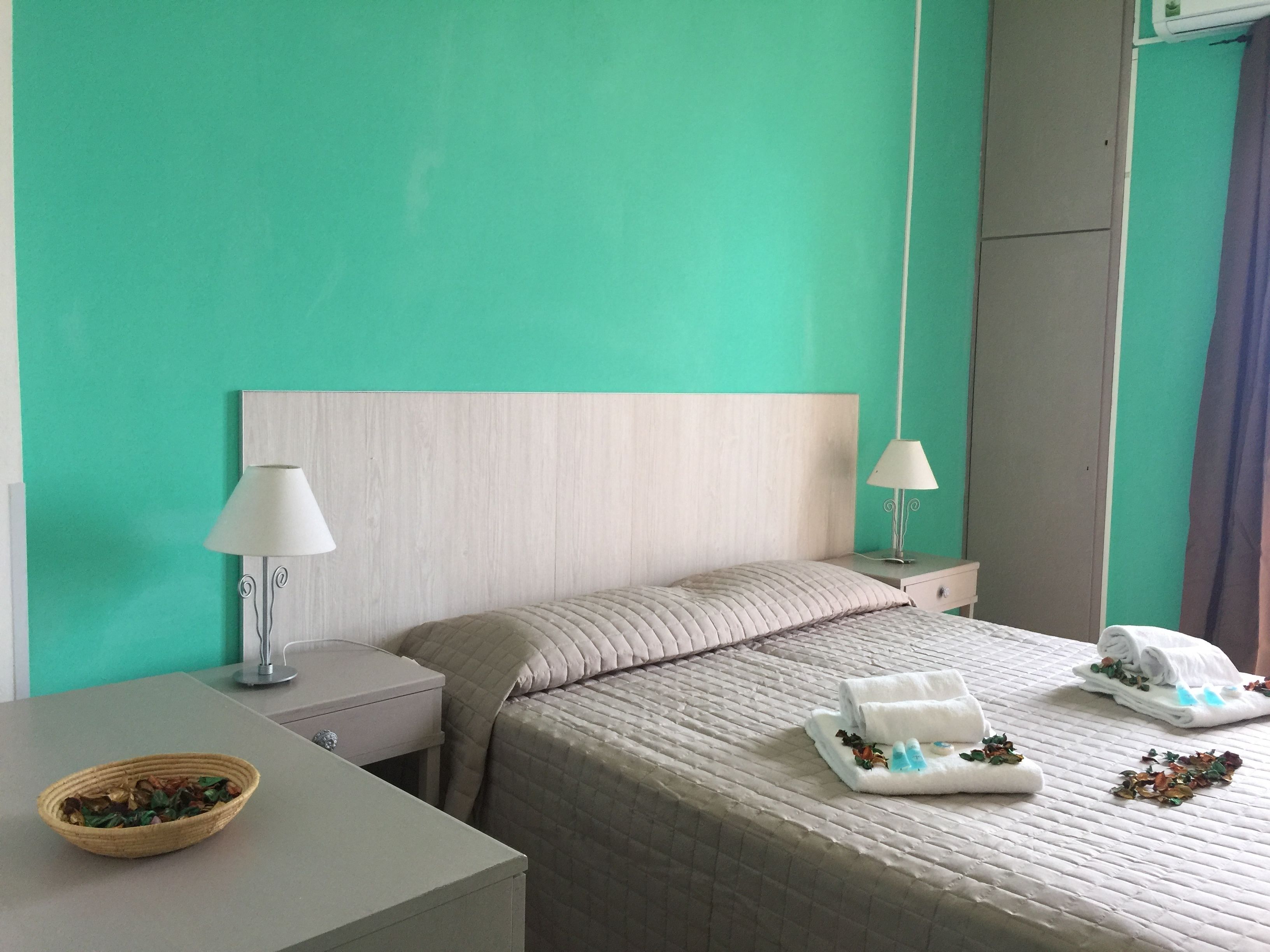 A hotel room with a bed, two bedside tables, a lamp on each table, a bowl of dried flowers on the left table, and a folded towel with a decorative item on the bed.
