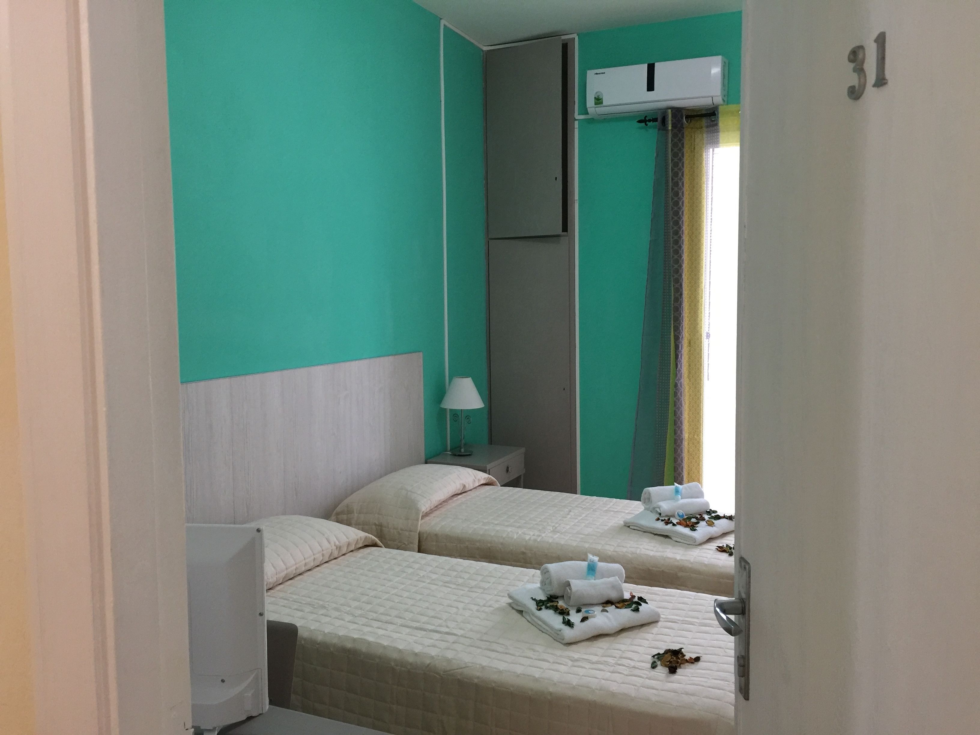 A hotel room with two beds, a bedside table with a lamp, an air conditioning unit, and a curtain.