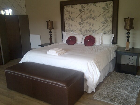 A neatly made bed with white linens, two red decorative pillows, a brown ottoman, and two bedside lamps on nightstands.