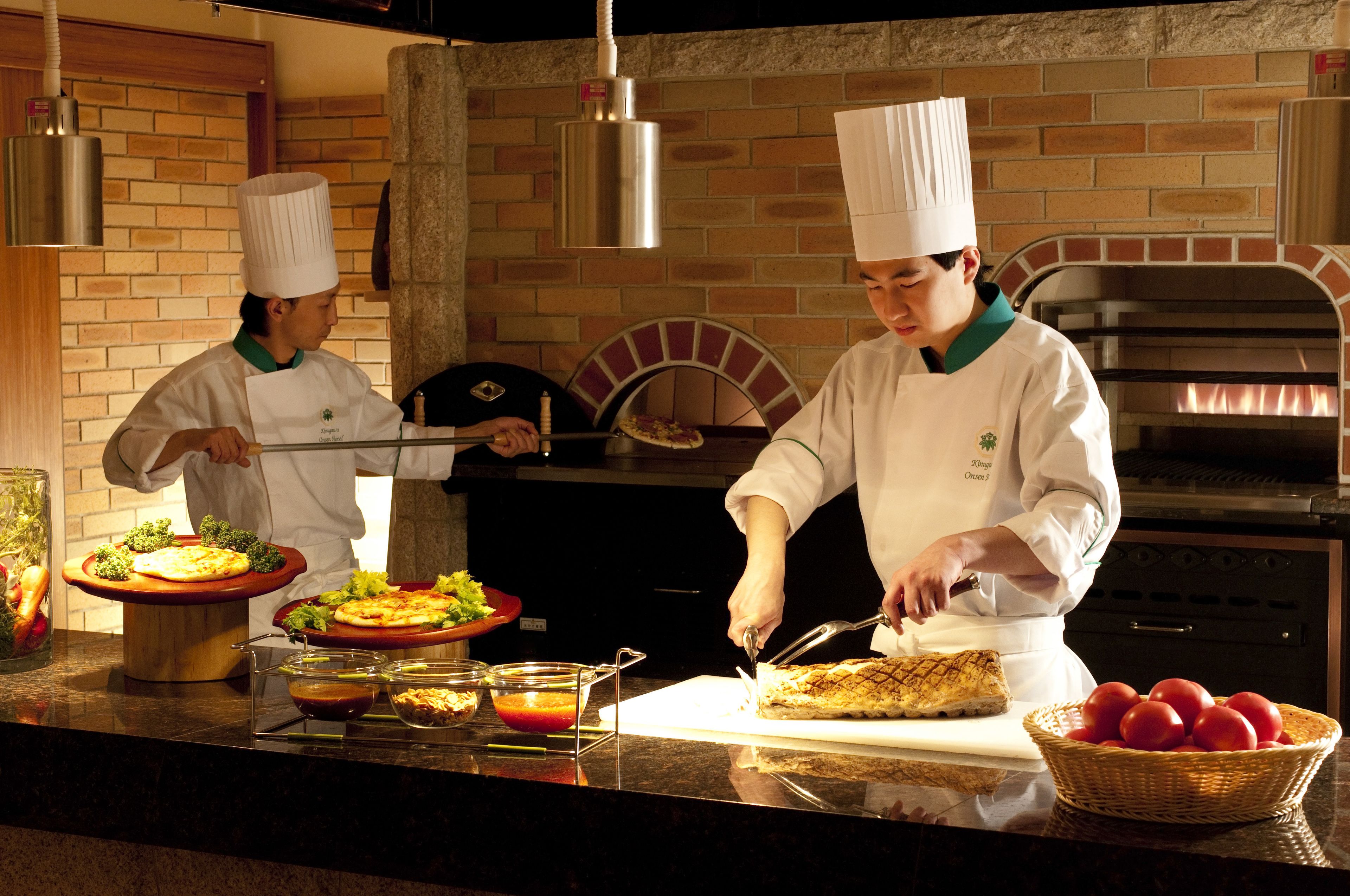 Chefs preparing food in a kitchen with brick walls, a brick oven, and a buffet with various dishes.