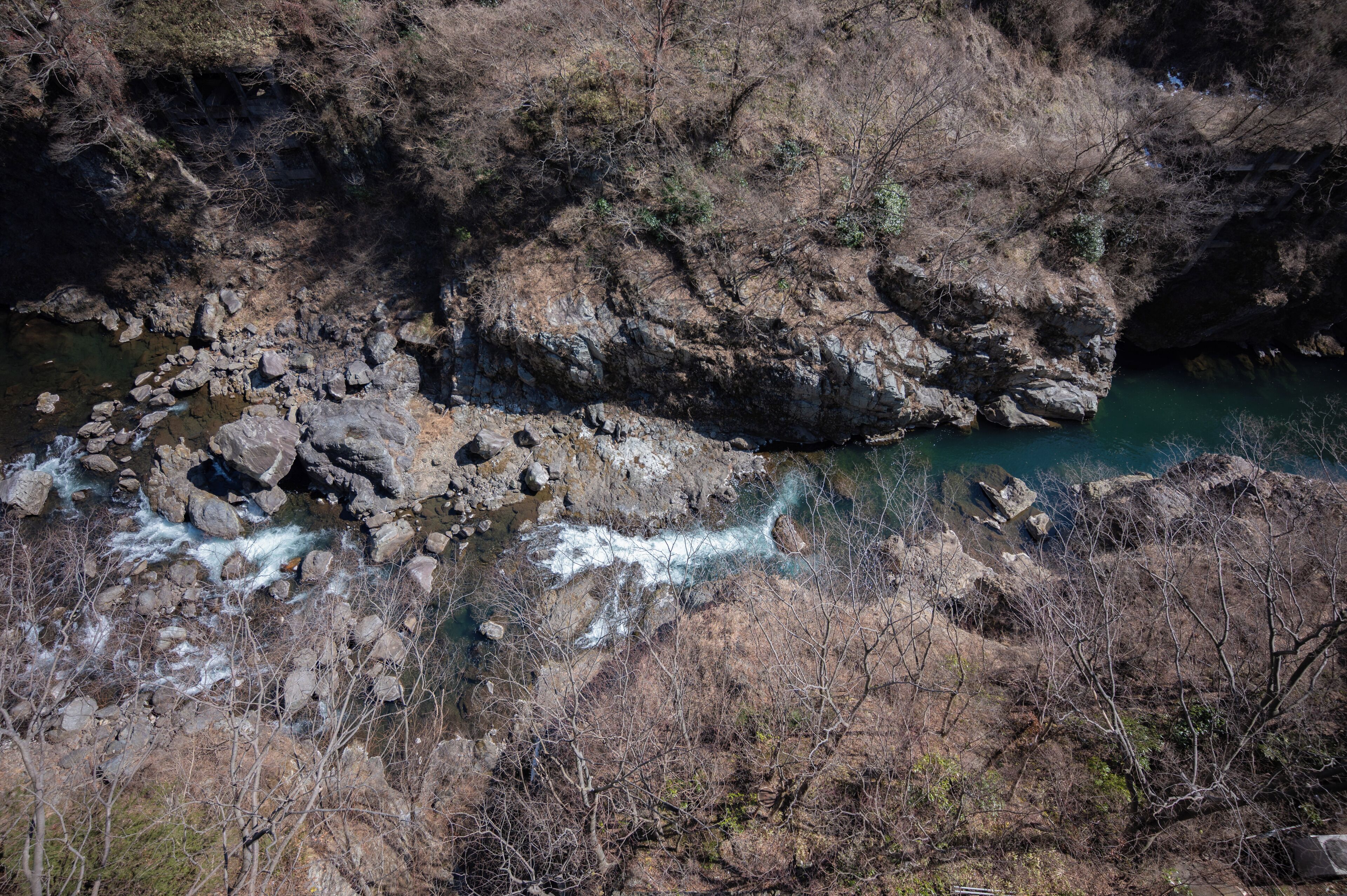 A rocky riverbed with clear water flowing through it, surrounded by dry, leafless trees.