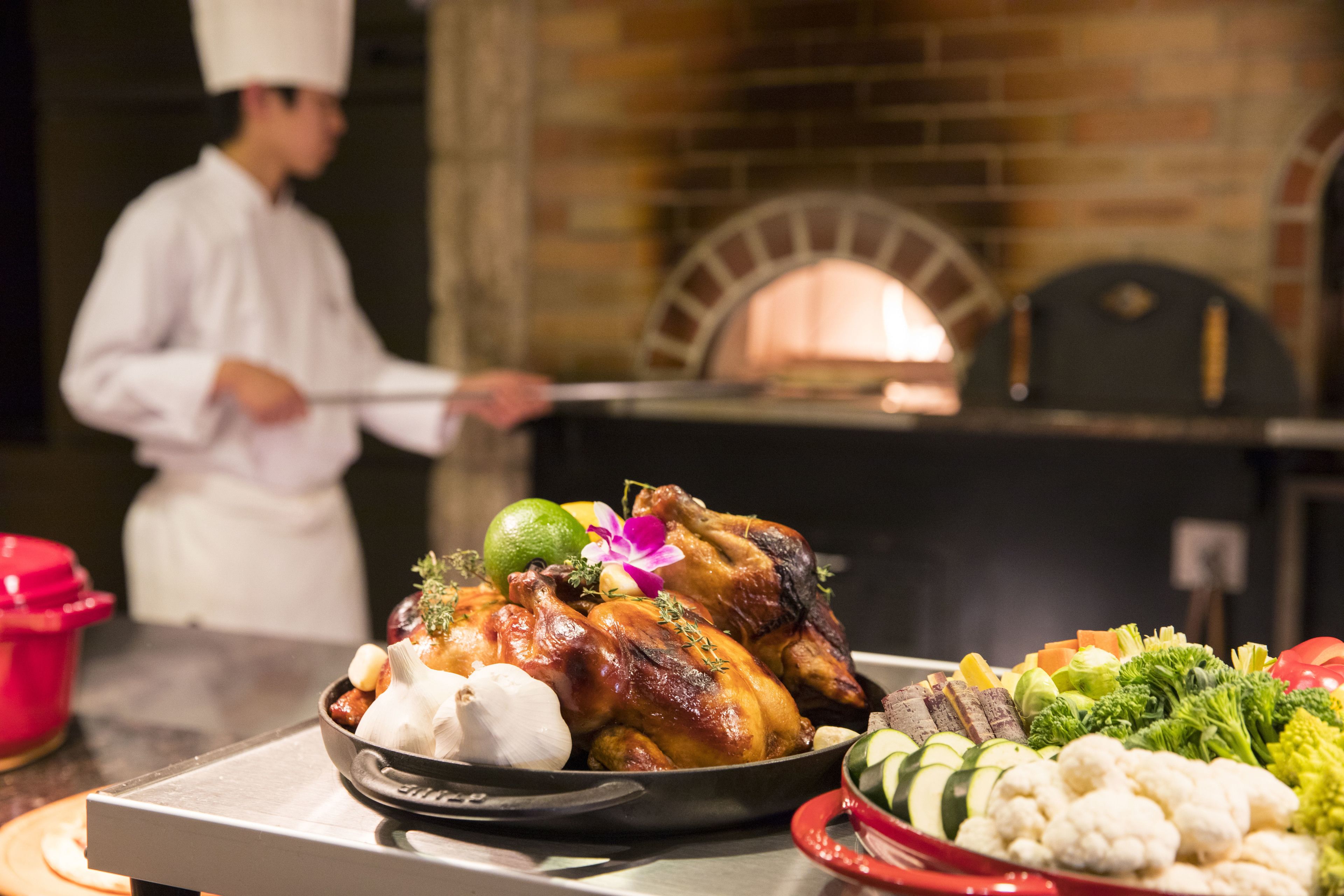 A chef in a white uniform preparing food in a kitchen with a brick oven in the background.