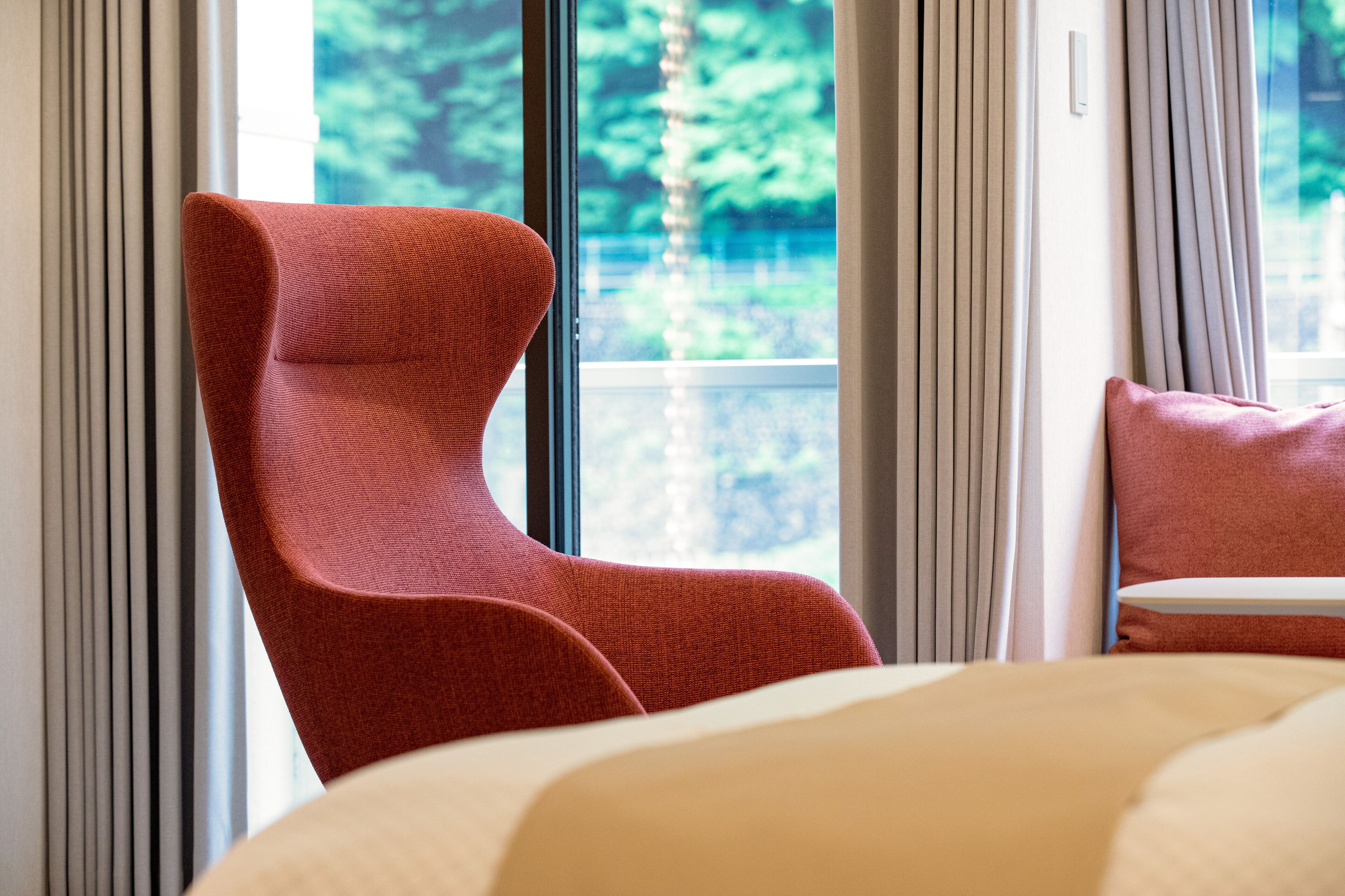 Una habitación de hotel moderna con una cama grande, un sillón tapizado de rojo y vistas a zonas verdes por la ventana.