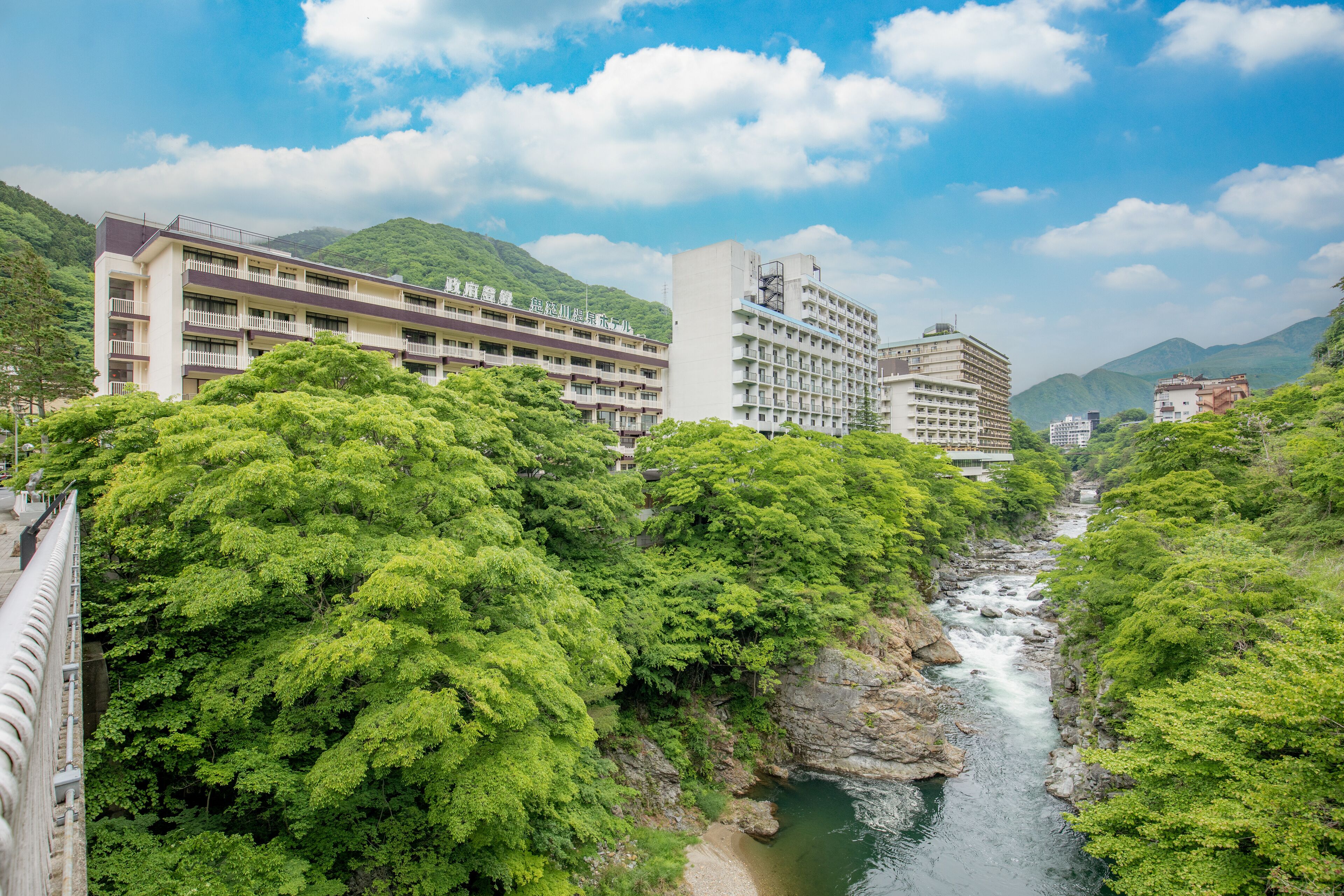 A multi-story hotel with balconies, a river, and lush greenery in the background.