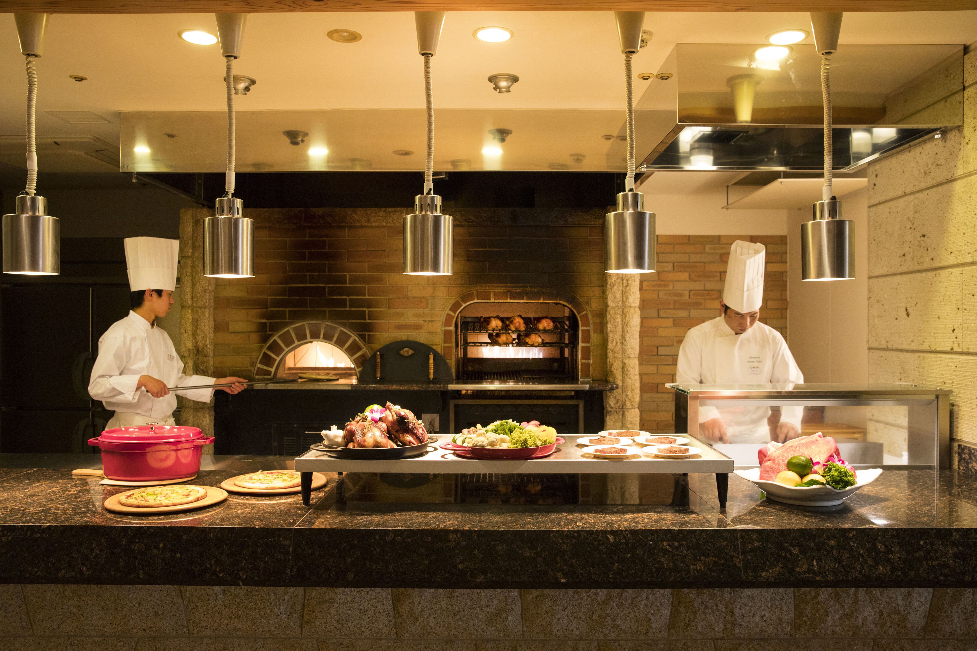A professional kitchen with chefs preparing food, a brick oven, and various dishes on the counter.