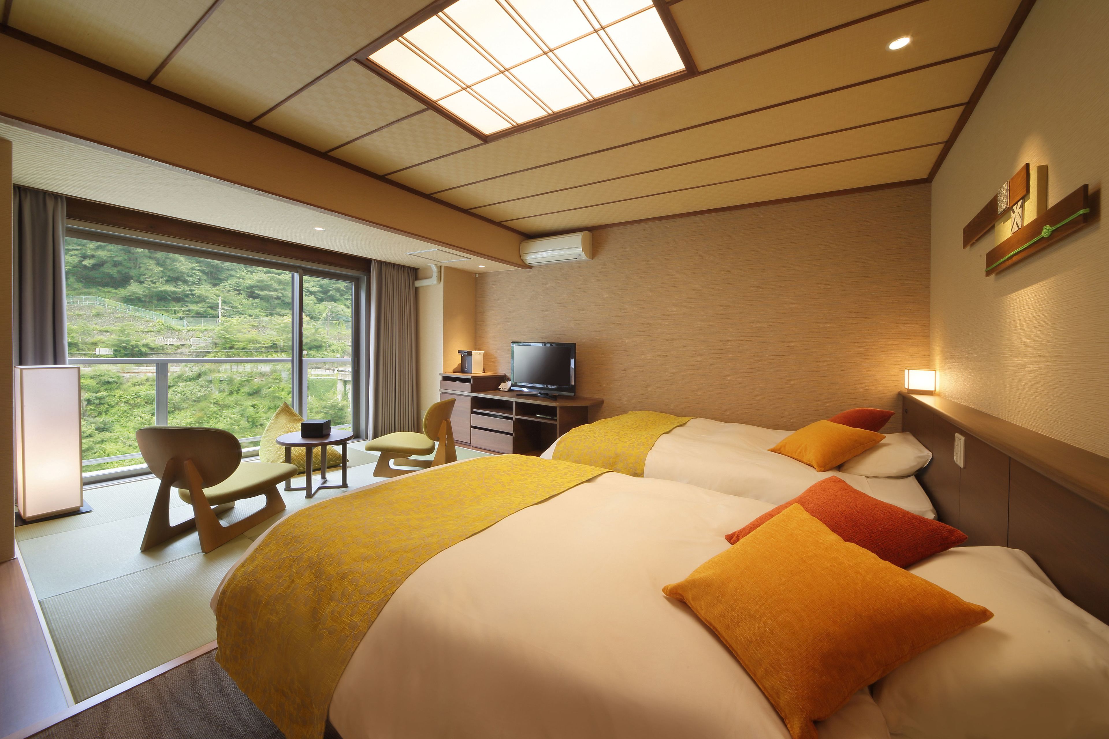 A hotel room with two beds, a TV, a desk, and a view of greenery outside.