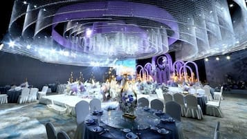 A banquet hall with round tables, chairs, and a central stage with floral arrangements.