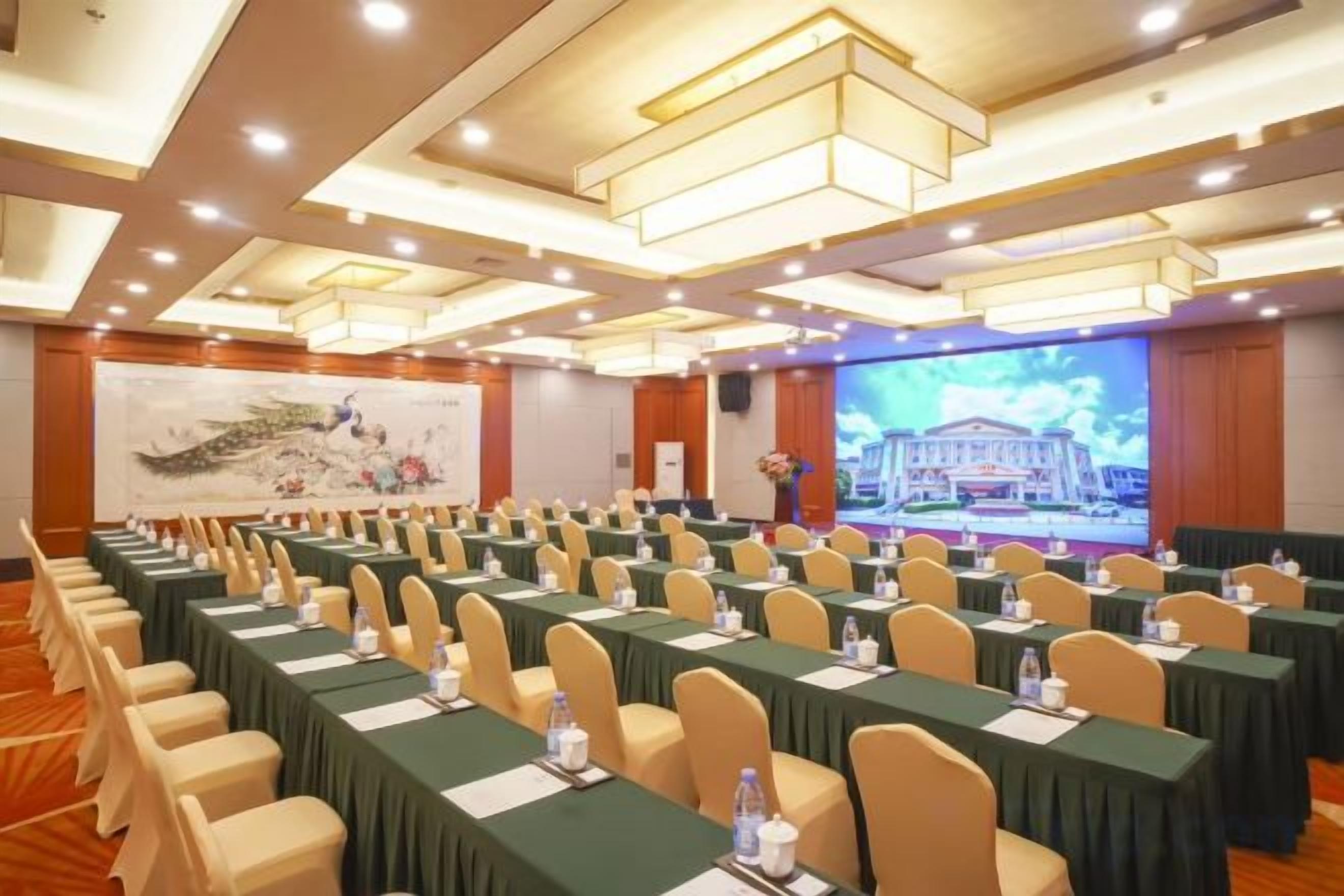 Un salón de conferencias con filas de mesas y sillas, una pantalla grande y un escenario con un atril.