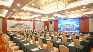 Un salón de conferencias con filas de mesas y sillas, una pantalla grande y un escenario con un atril.