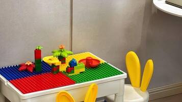 Una mesa blanca con un tapete de juego LEGO y una silla de juguete con detalles azules y amarillos.