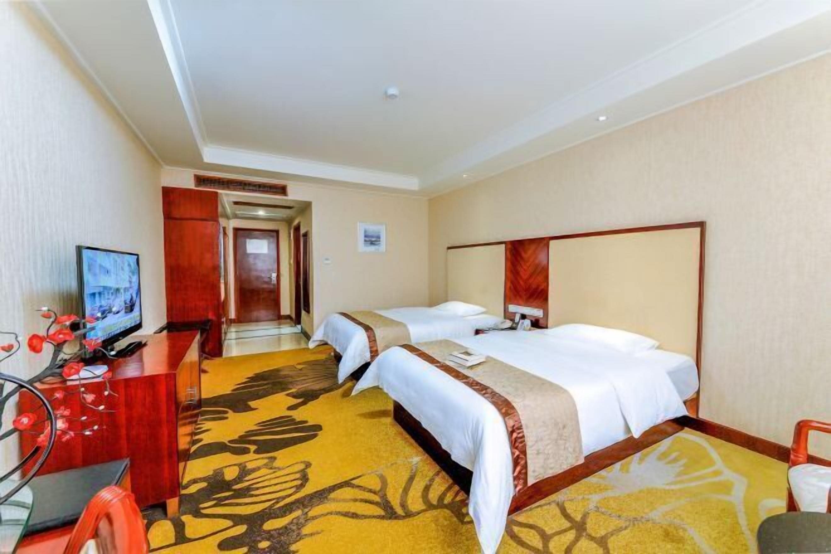 A hotel room with two beds, a desk with a television, and a carpet with a floral pattern.
