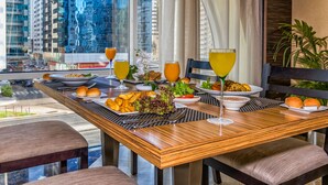 Une table à manger dressée avec différents plats et boissons, une vue sur la ville à travers de grandes fenêtres et un aménagement intérieur moderne.